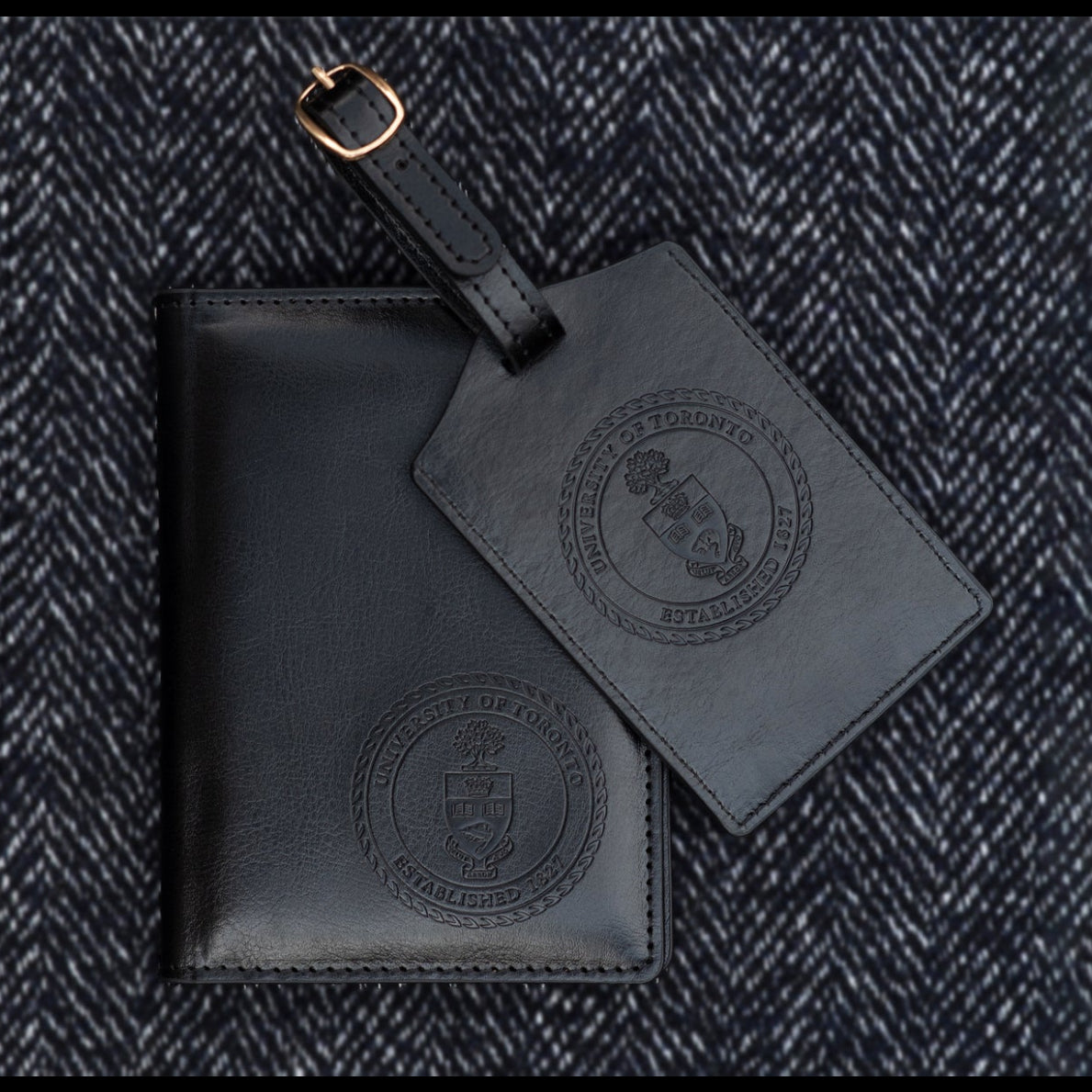 A black leather luggage tag and passport holder with debossed logos on a textured dark fabric background